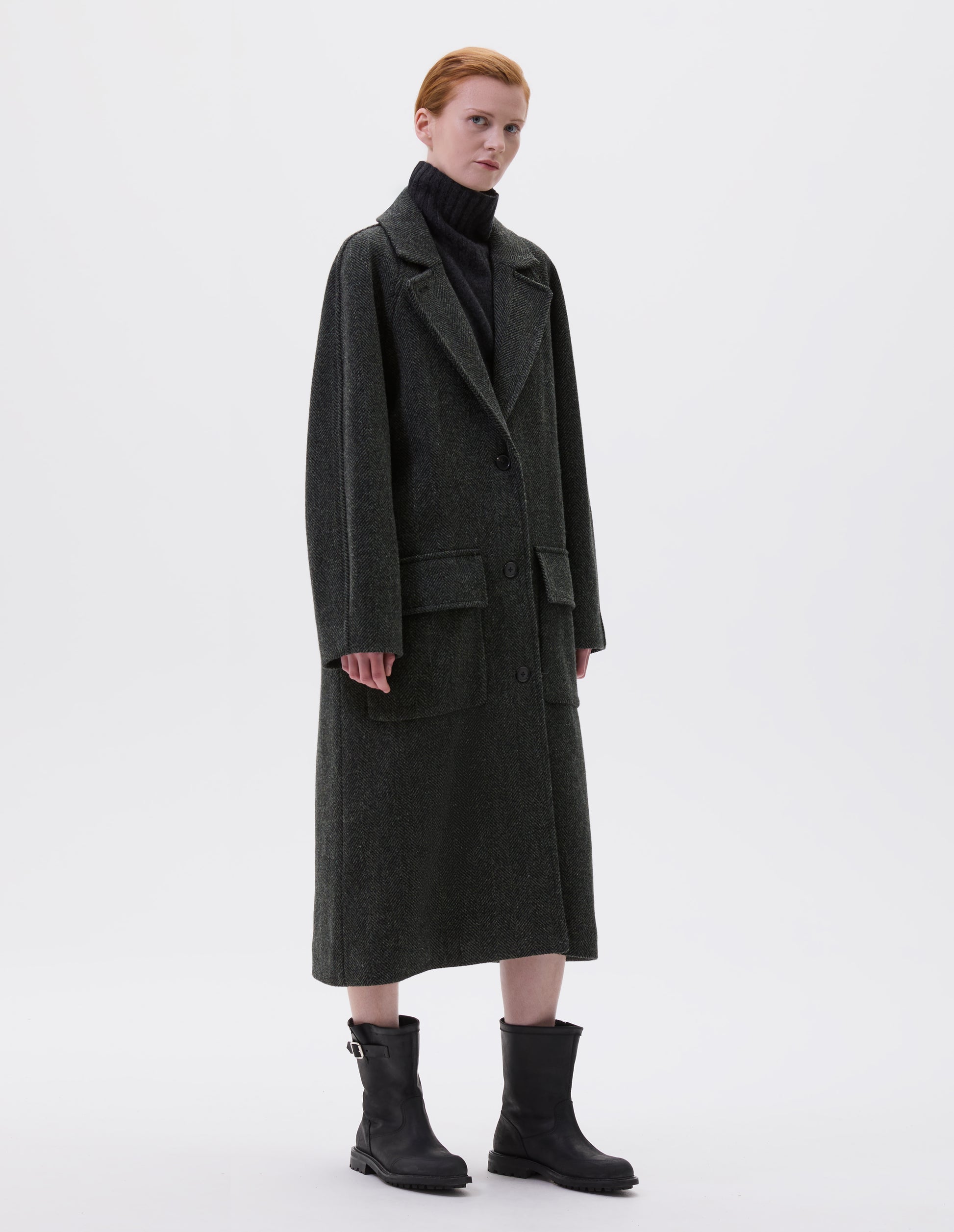 Full body image, side view of model wearing green and black herringbone wool longline overcoat with raglan sleeve, button fastening lapel and deep flap patch pockets. Styled with grey high neck jumper and black leather boots.