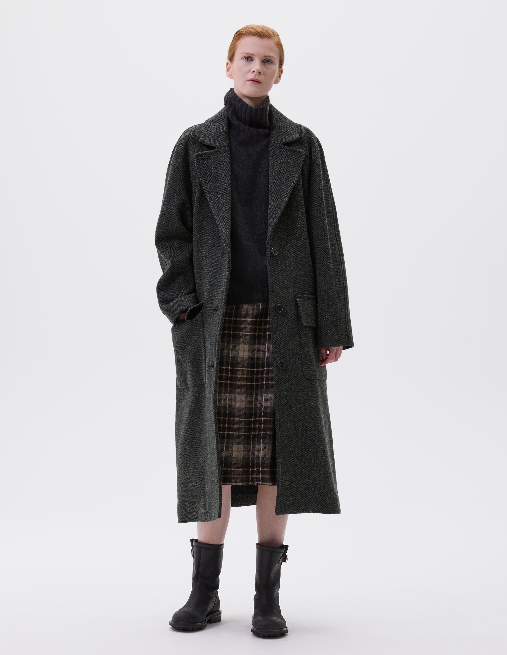 Full body image, front view of model wearing green and black herringbone wool longline overcoat with raglan sleeve, button fastening lapel and deep flap patch pockets. Styled with grey high neck jumper, brown and beige checked skirt and black leather boots.