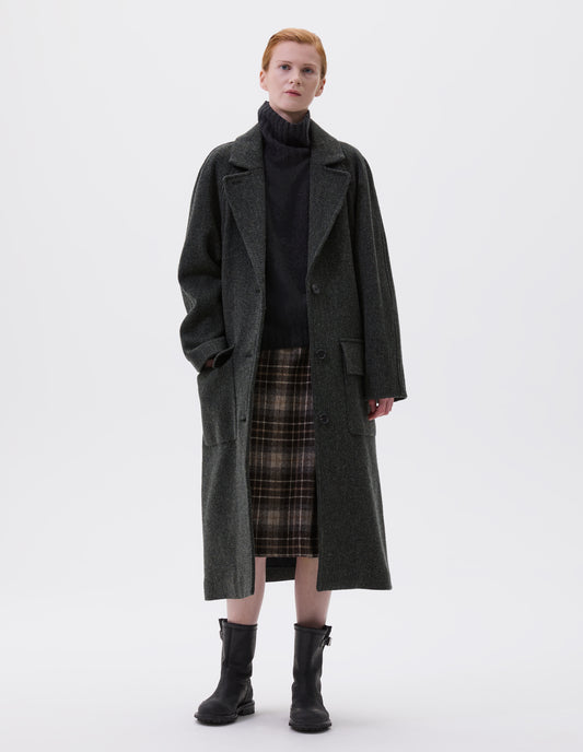 Full body image, front view of model wearing green and black herringbone wool longline overcoat with raglan sleeve, button fastening lapel and deep flap patch pockets. Styled with grey high neck jumper, brown and beige checked skirt and black leather boots.