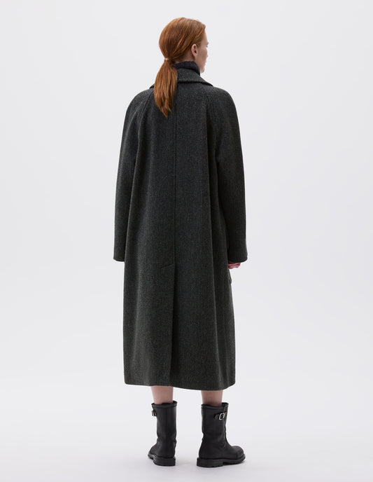Full body image, back view of model wearing green and black herringbone wool longline overcoat with raglan sleeve and back centre vent. Styled with black leather boots. 