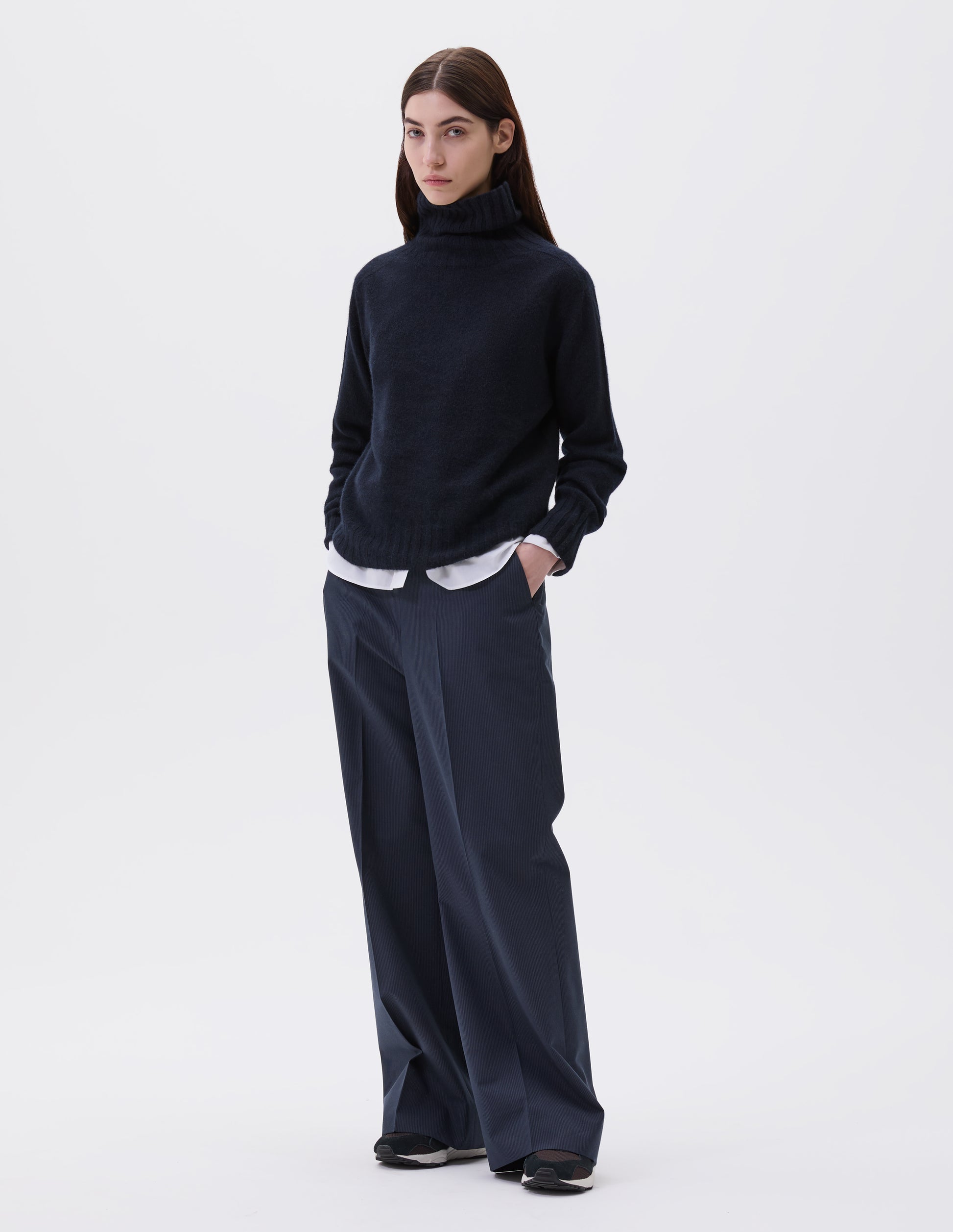 Full body image, front view of model wearing dark navy blue cashmere roll neck jumper with contrast ribbing on neck, cuffs and hem. Styled with white cotton shirt, navy blue pinstripe trouser and black and brown trainers.