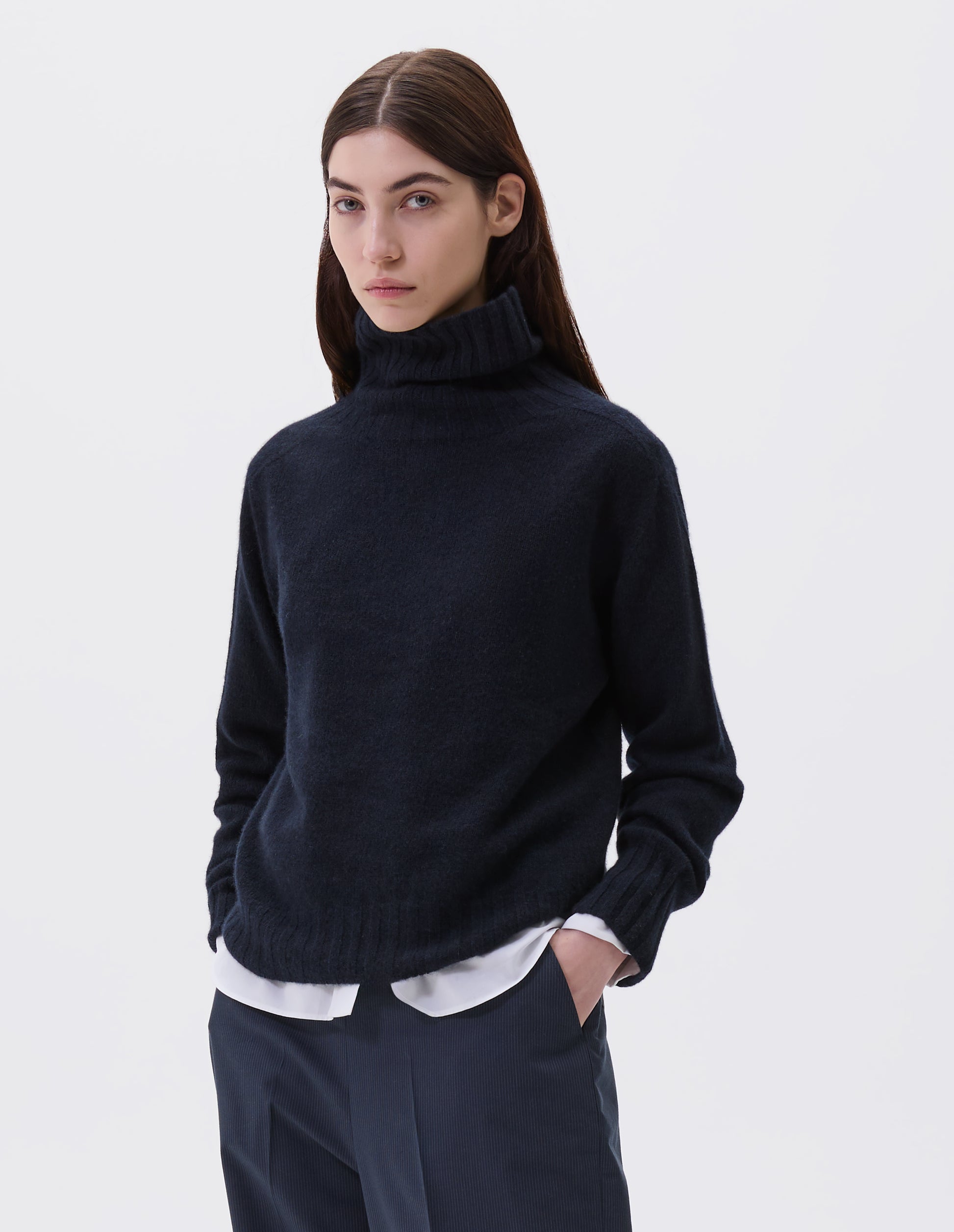 Cropped body image, front view of model wearing dark navy blue cashmere roll neck jumper with contrast ribbing on neck, cuffs and hem. Styled with white cotton shirt and navy blue pinstripe trousers.