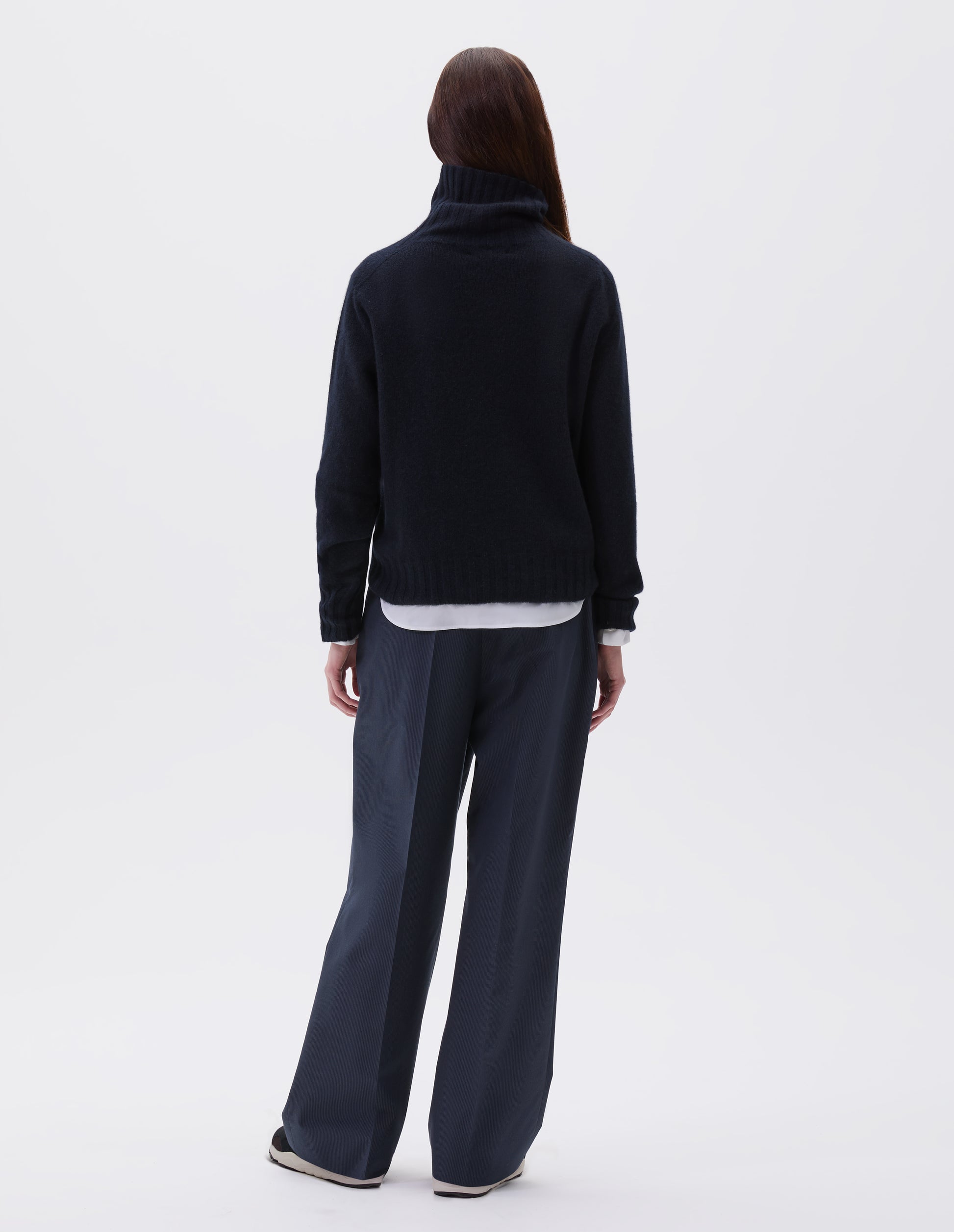 Full body image, back view of model wearing dark navy blue cashmere roll neck jumper with contrast ribbing on neck, cuffs and hem. Styled with white cotton shirt, navy blue pinstripe trouser and black and brown trainers.