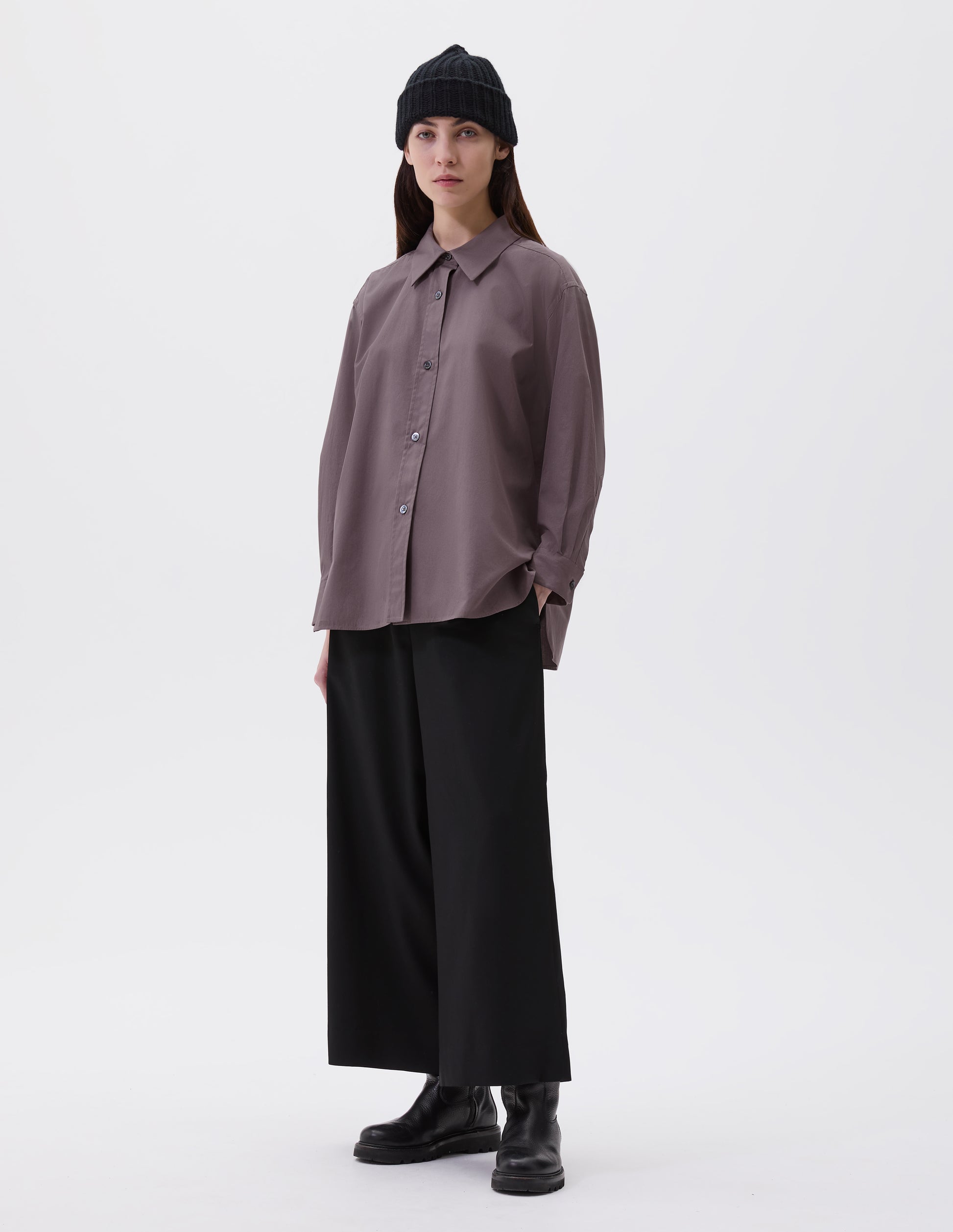 Full body image, front view of model wearing relaxed brown shirt with raised, stitch-through placket with mother of pearl buttons and A line hem. Styled with black beanie, black culottes and black leather boots.