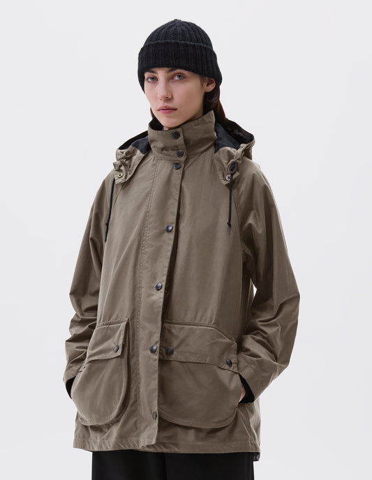 Cropped image, front view of model wearing oversized dark beige parka with stand collar and hood with wire adjustable peak, raglan sleeve, press stud fastening storm flap, large bellows pockets with press stud fastening envelope closure and concealed side pockets. Styled with black beanie and black culottes. 