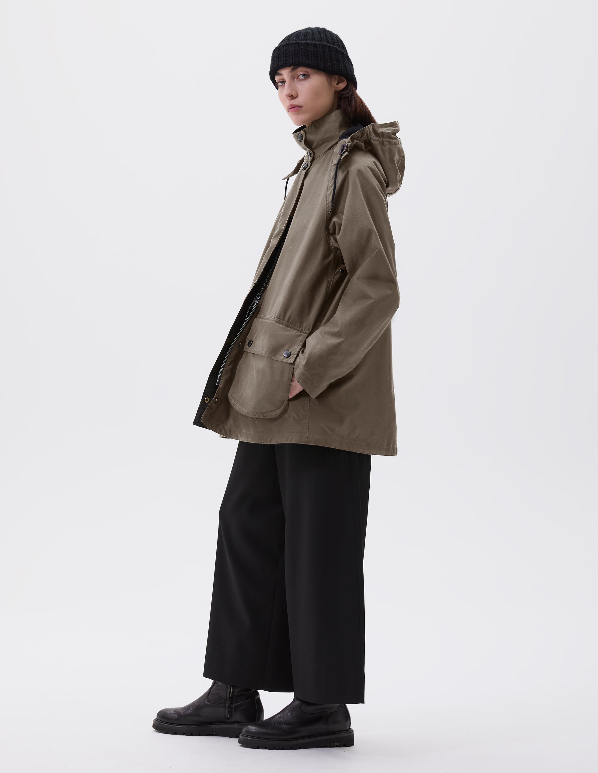 Full body image, side view of model wearing oversized dark beige parka with stand collar and hood with wire adjustable peak, two way zip, raglan sleeve, press stud fastening storm flap and large bellows pockets with press stud fastening envelope closure with concealed side pockets. Styled with black beanie, black culottes and black leather boots.
