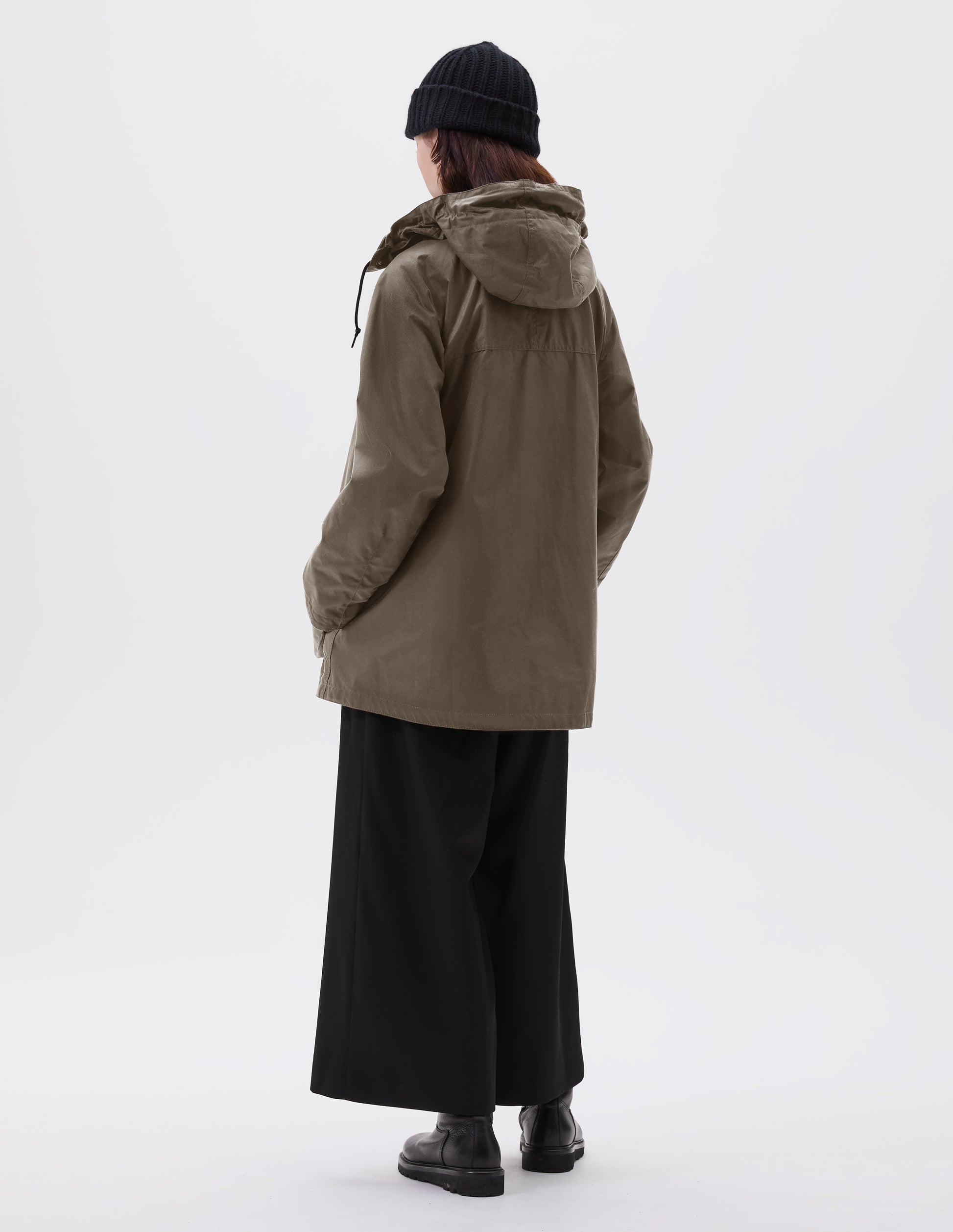 Full body image, back view of model wearing oversized dark beige parka with adjustable hood and raglan sleeves. Styled with black beanie, black culottes and black leather boots.