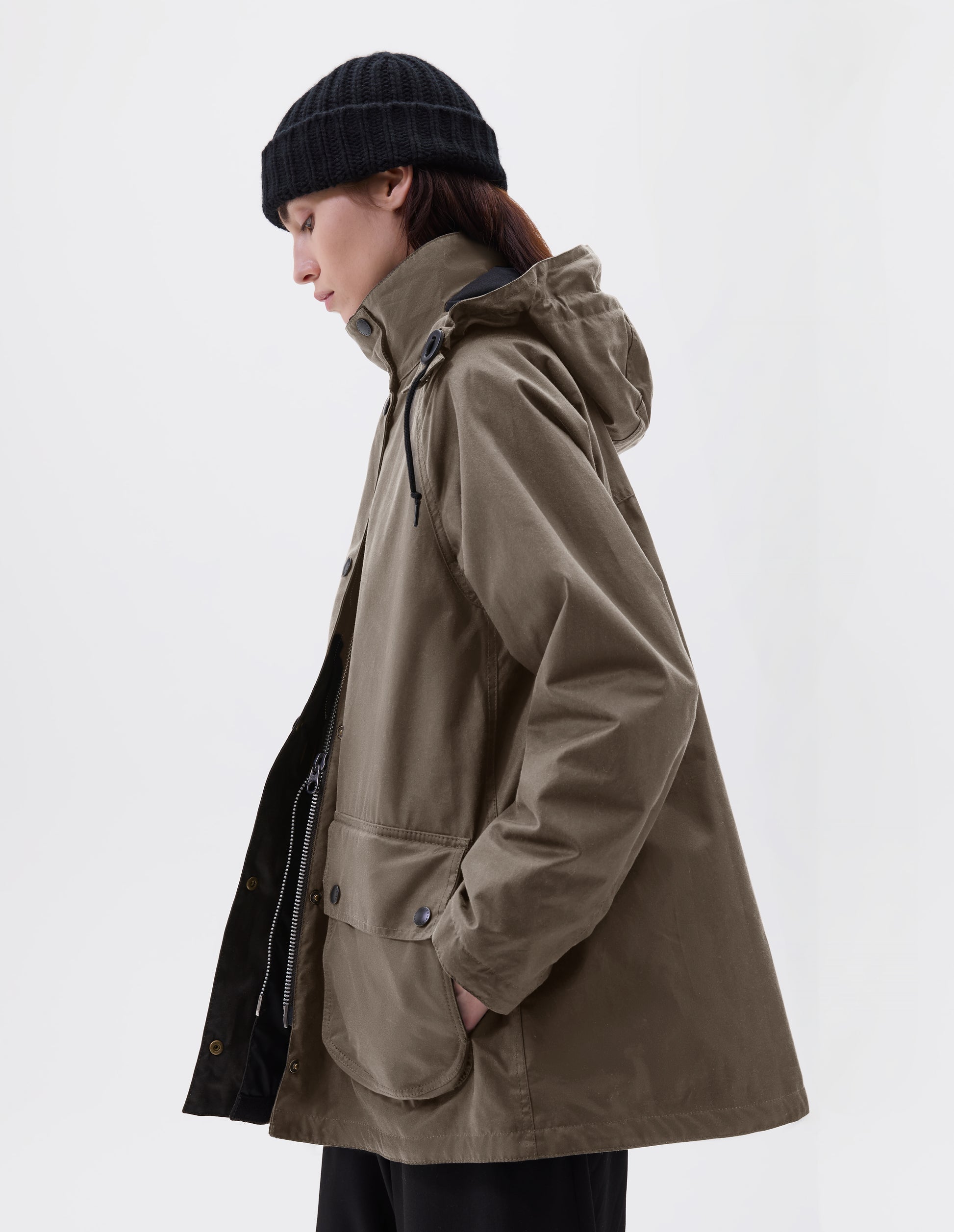 Cropped image, side view of model wearing oversized dark beige parka with stand collar and hood with wire adjustable peak, two way zip, raglan sleeve, press stud fastening storm flap, large bellows pockets with press stud fastening envelope closure and concealed side pockets. Styled with black beanie and black culottes. 