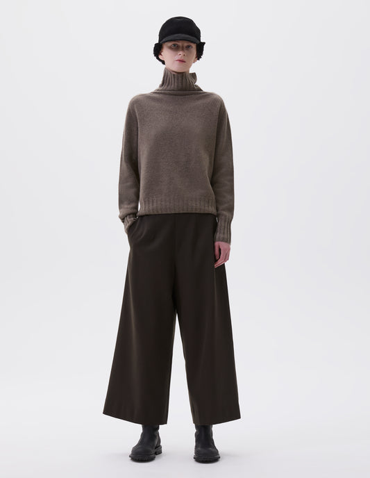 Full body image, front view of model wearing beige high neck cashmere jumper, with saddle sleeve and contrasting wide rib on neck, cuffs and hem. Styled with black shearling cap, black culottes and black leather boots.
