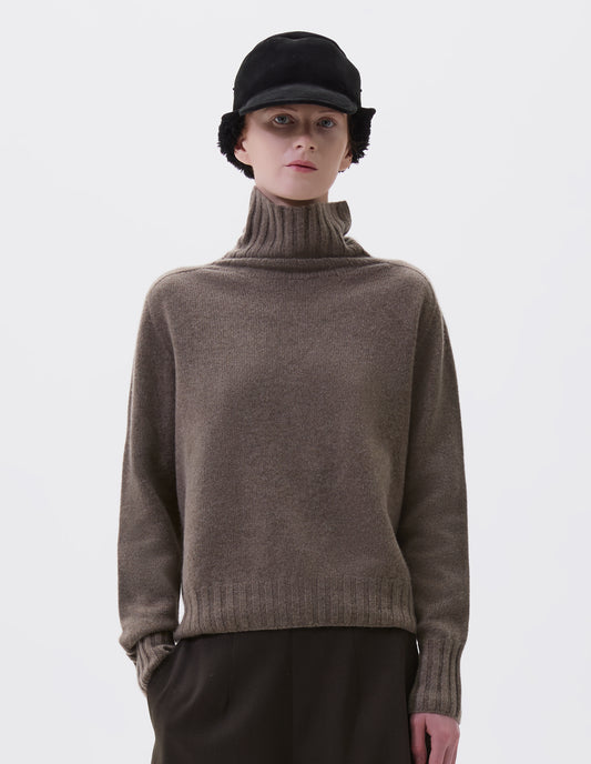 Cropped body image, front view of model wearing beige high neck cashmere jumper, with saddle sleeve and contrasting wide rib on neck, cuffs and hem. Styled with black shearling cap and black culottes.