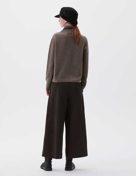 Full body image, back view of model wearing beige high neck cashmere jumper, with saddle sleeve and contrasting wide rib on neck, cuffs and hem. Styled with black shearling cap, black culottes and black leather boots.