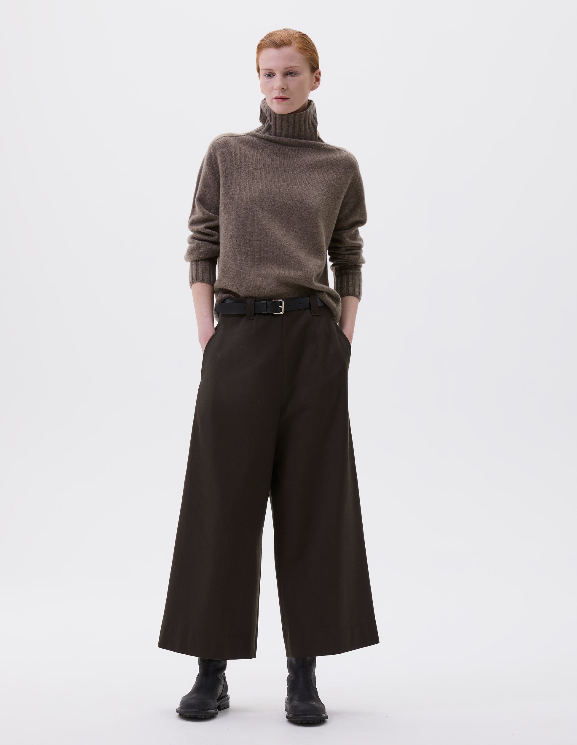 Full body image, front view of model wearing brownish green wool wide leg culottes, with belt loops, side pockets and concealed zip fastening. Styled with beige turtleneck jumper, black leather belt and black leather boots.