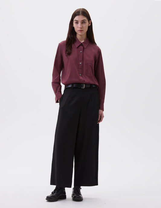 Full body image, front view of model wearing dark red shirt with raised placket and mother of pearl buttons, patch pocket on chest, styled with black leather belt, black trousers and black socks with black brogues.