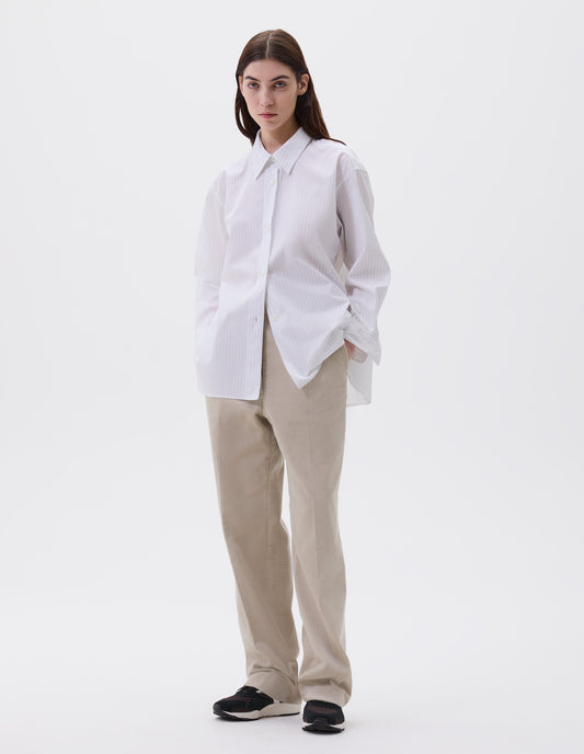 Full body image, front view of model wearing relaxed white and grey striped shirt with raised, stitch-through placket with mother of pearl buttons and A line hem. Styled with beige trousers and black, brown and white trainers.