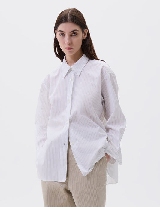 Cropped image, front view of model wearing relaxed white and grey striped shirt with raised, stitch-through placket with mother of pearl buttons and A line hem. Styled with beige trousers.