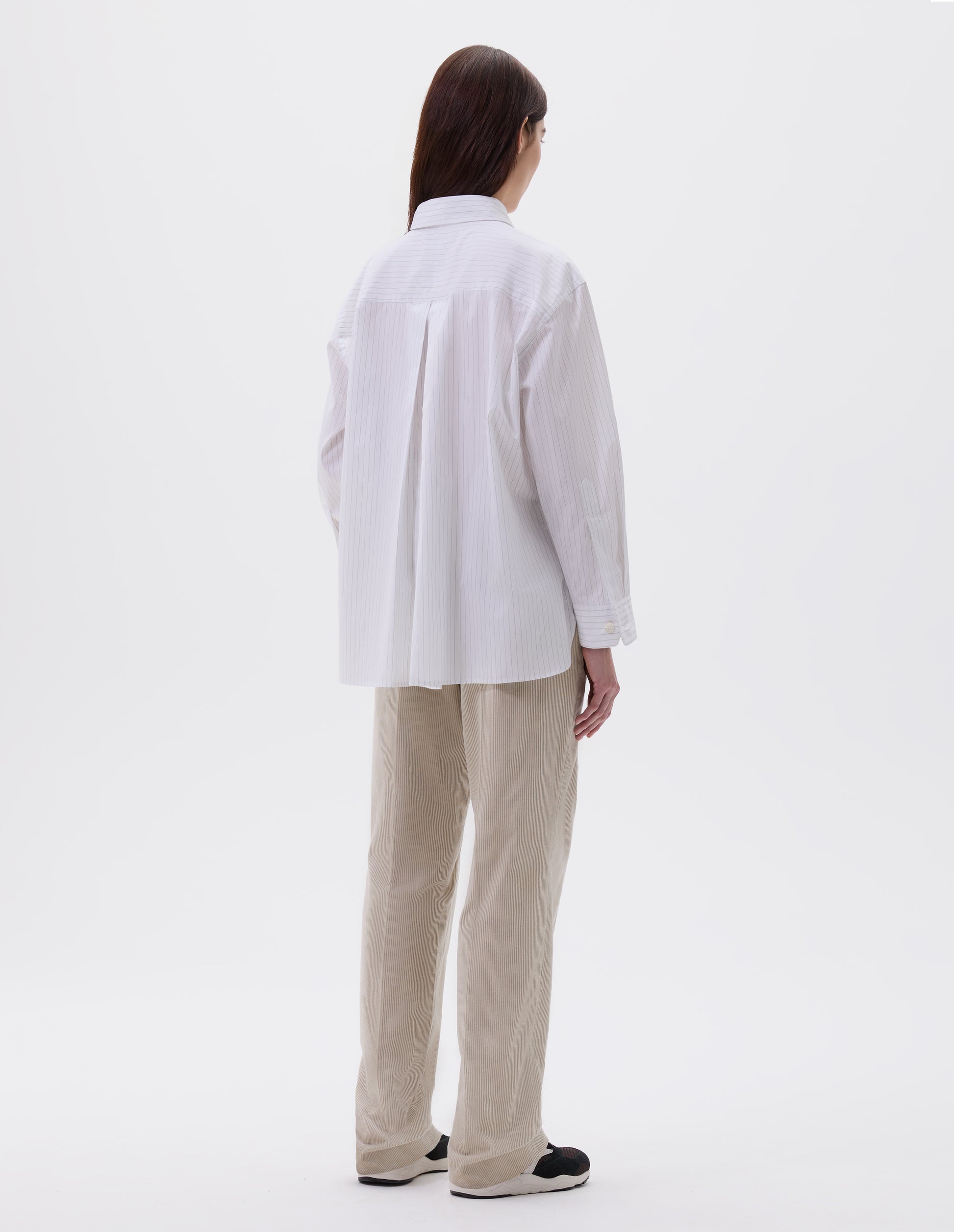 Full body image, back view of model wearing relaxed white and grey striped shirt with A line hem, split panel yoke with deep, inverted centre back pleat and mother of pearl buttons on cuffs. Styled with beige trousers and black, brown and white trainers.