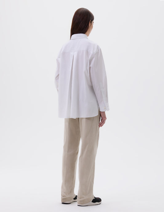 Full body image, back view of model wearing relaxed white and grey striped shirt with A line hem, split panel yoke with deep, inverted centre back pleat and mother of pearl buttons on cuffs. Styled with beige trousers and black, brown and white trainers.