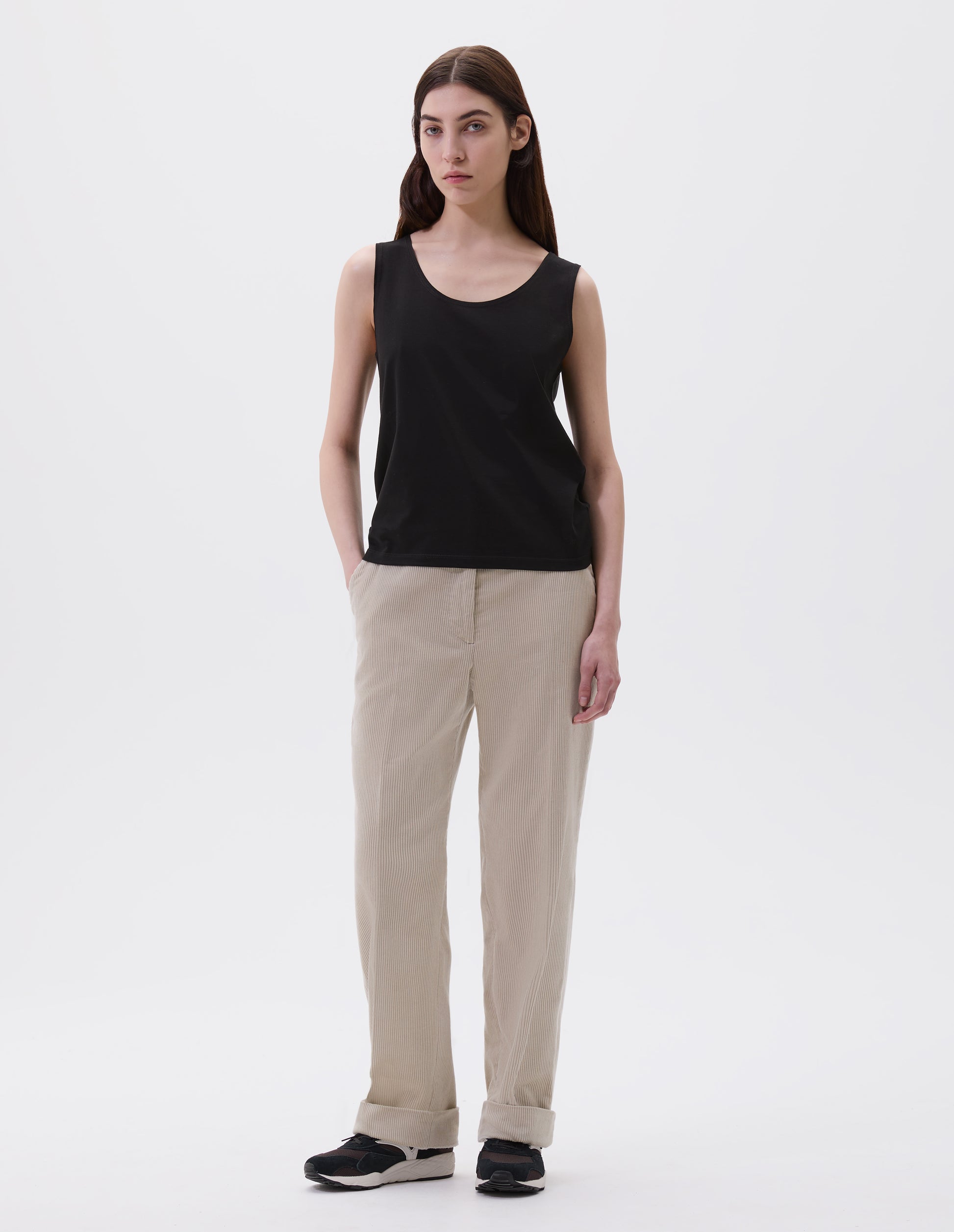 Full body image, front view of model wearing black cotton sleeveless vest with scoop neck. Styled with beige corduroy trousers and black and brown trainers. 