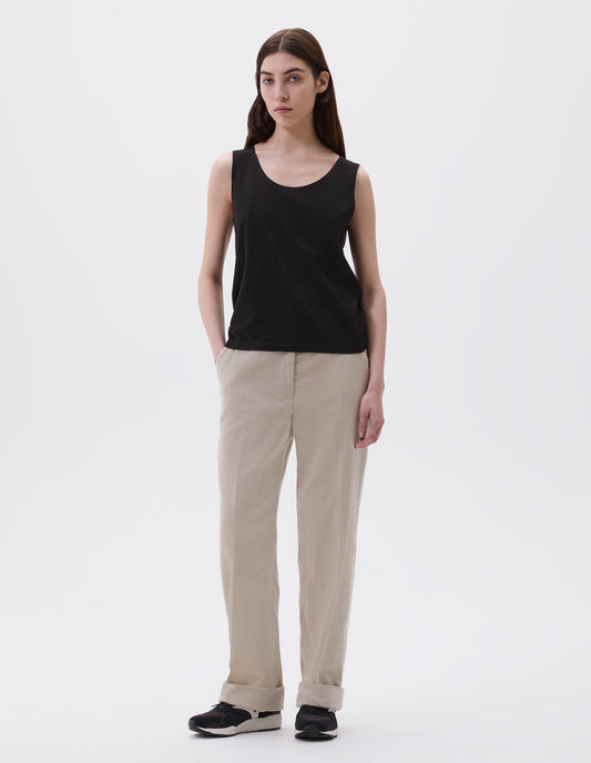 Full body image, front view of model wearing black cotton sleeveless vest with scoop neck. Styled with beige corduroy trousers and black and brown trainers. 
