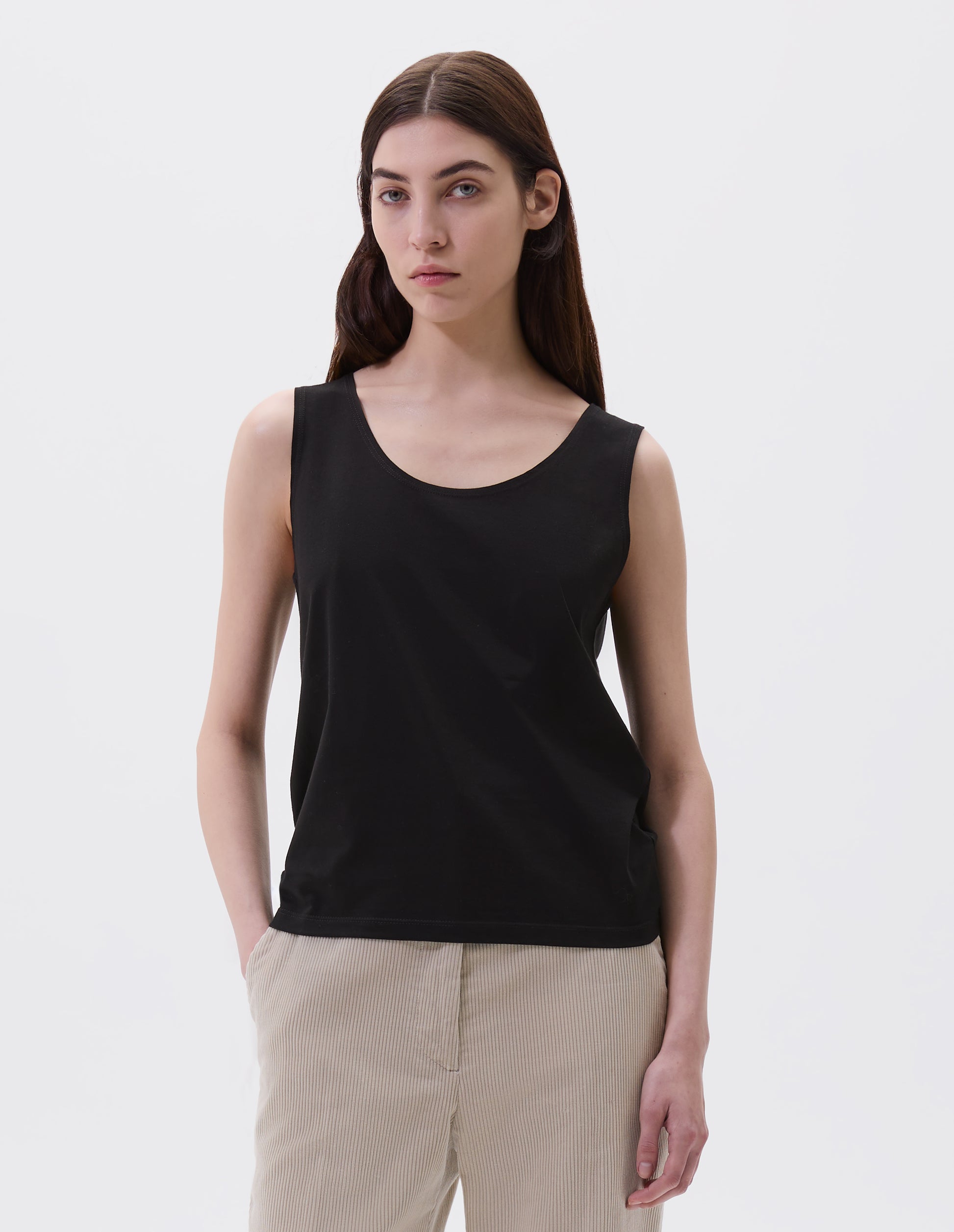 Cropped body image, front view of model wearing black cotton sleeveless vest with scoop neck. Styled with beige corduroy trousers.