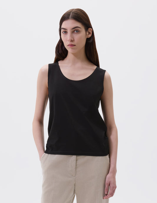 Cropped body image, front view of model wearing black cotton sleeveless vest with scoop neck. Styled with beige corduroy trousers.
