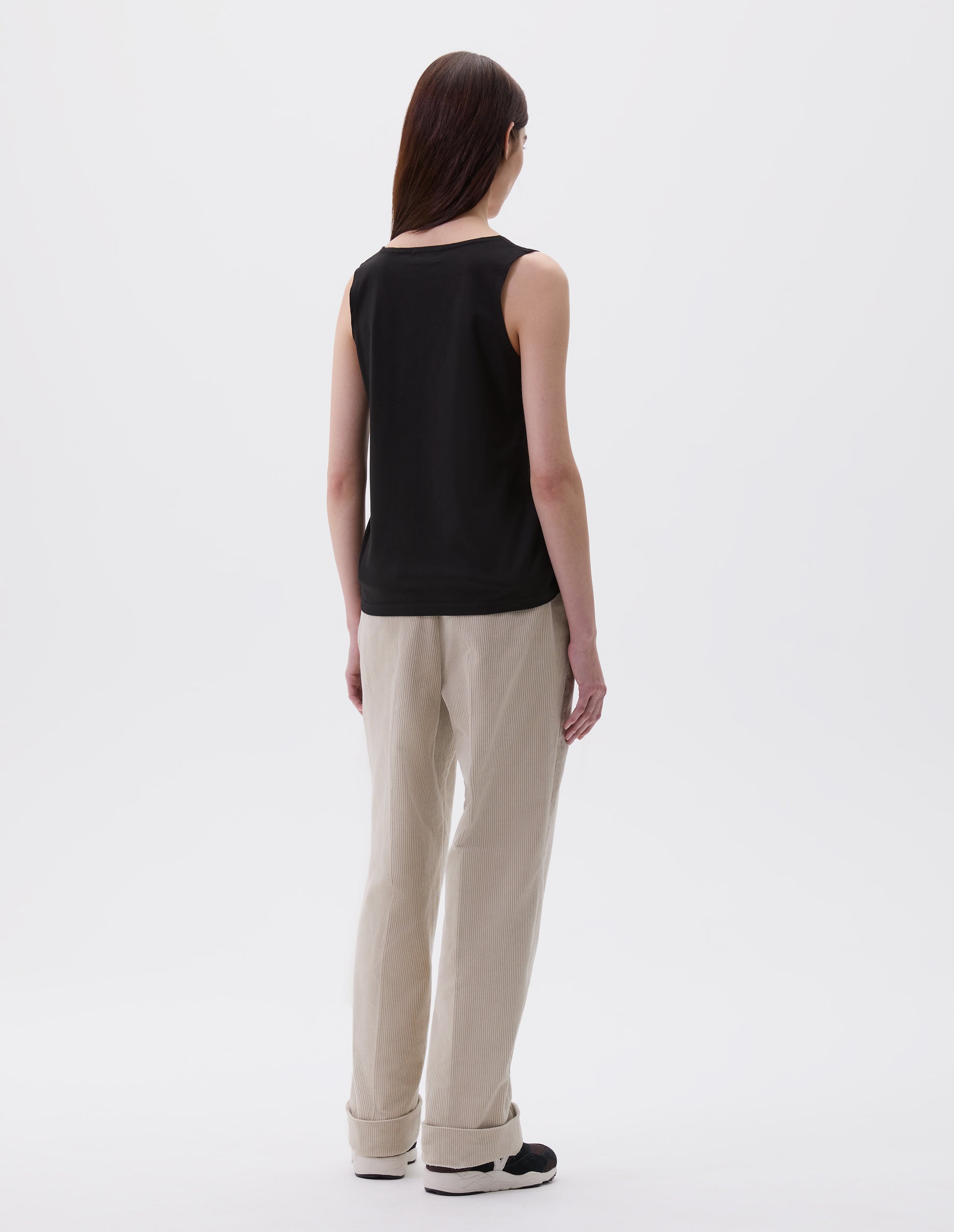 Full body image, back view of model wearing black cotton sleeveless vest. Styled with beige corduroy trousers and black and brown trainers. 