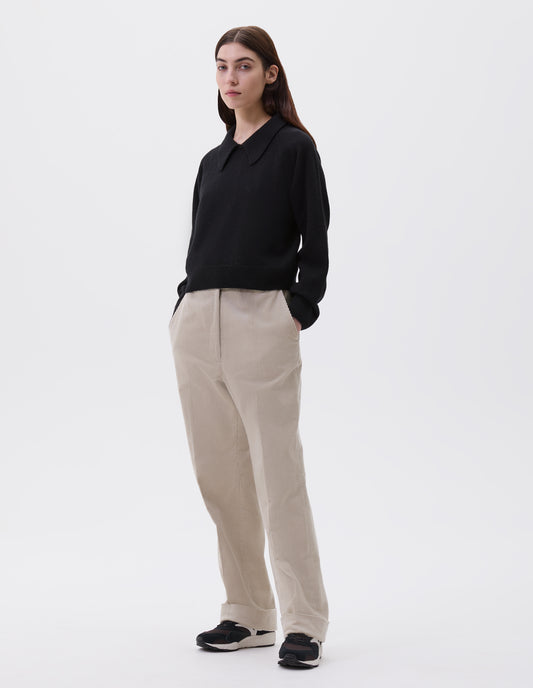Full body image, front view of model wearing black cropped collared jumper with contrast rib on cuffs and hem. Styled with beige trousers with side pockets and black, brown and white trainers.