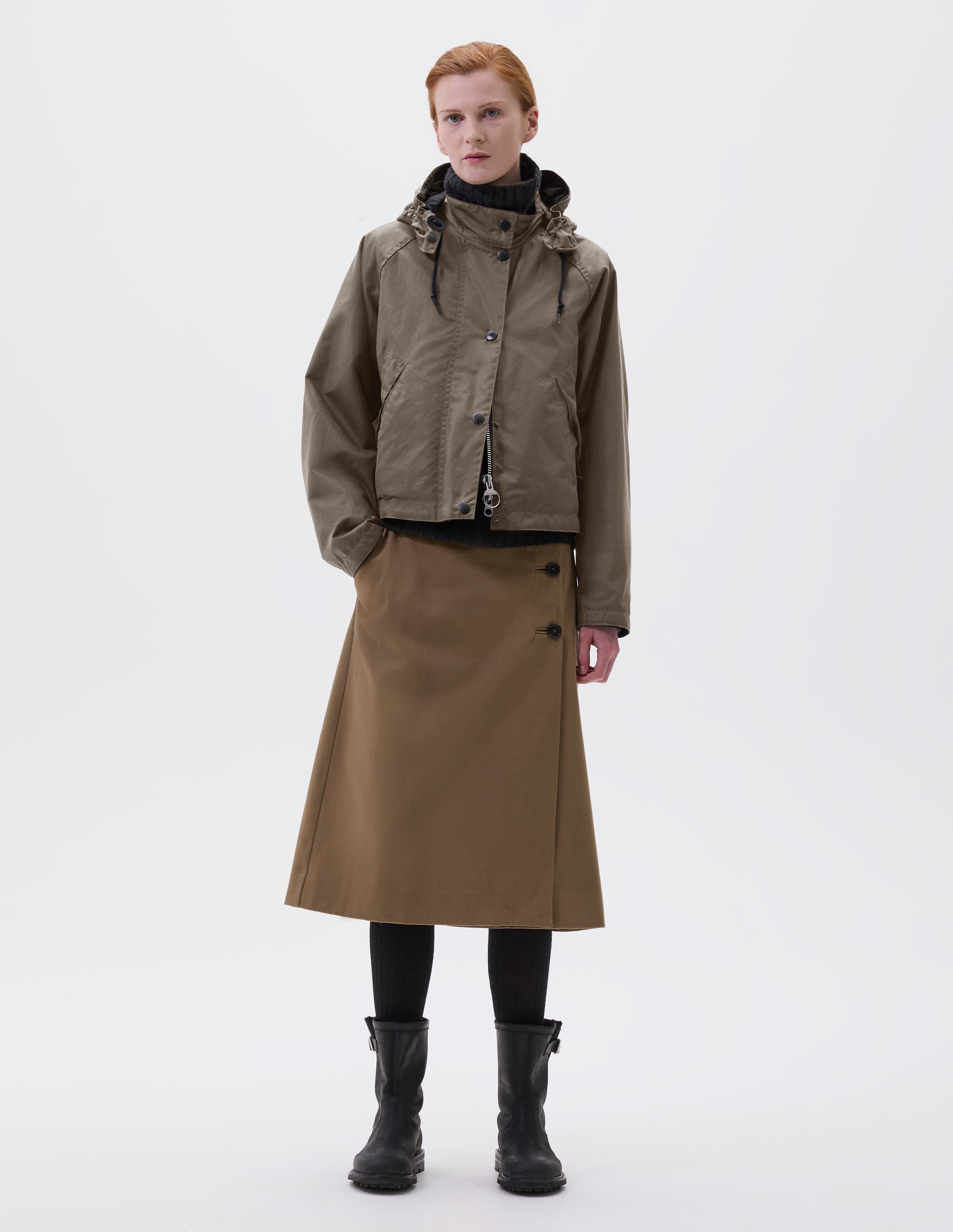 Full body image, front view of model wearing boxy dark beige jacket with stand collar and hood with wire adjustable peak, raglan sleeve, two-way zip fastening, press stud storm flap and wide storm welt side pockets. Styled over dark grey roll neck jumper with beige midi skirt, black tights and black biker boots.
