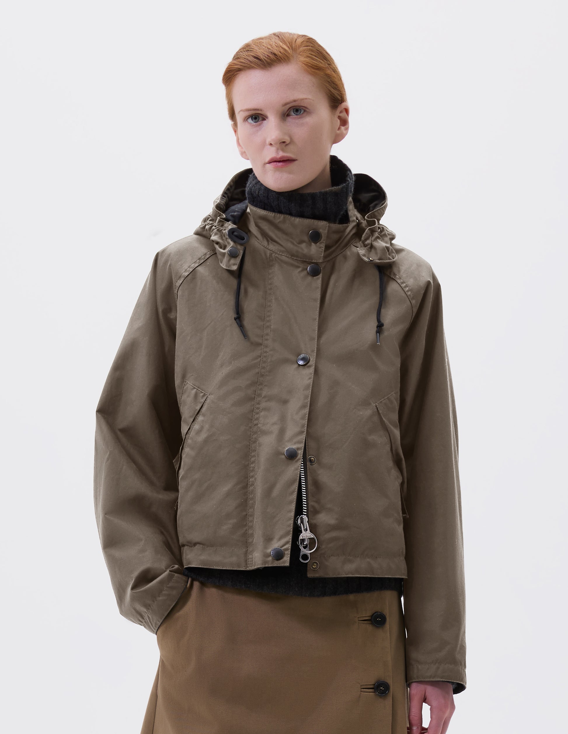 Cropped image, front view of model wearing boxy dark beige jacket with stand collar and hood with wire adjustable peak, raglan sleeve, two-way zip fastening, press stud storm flap and wide storm welt side pockets. Styled over dark grey roll neck jumper and beige midi skirt