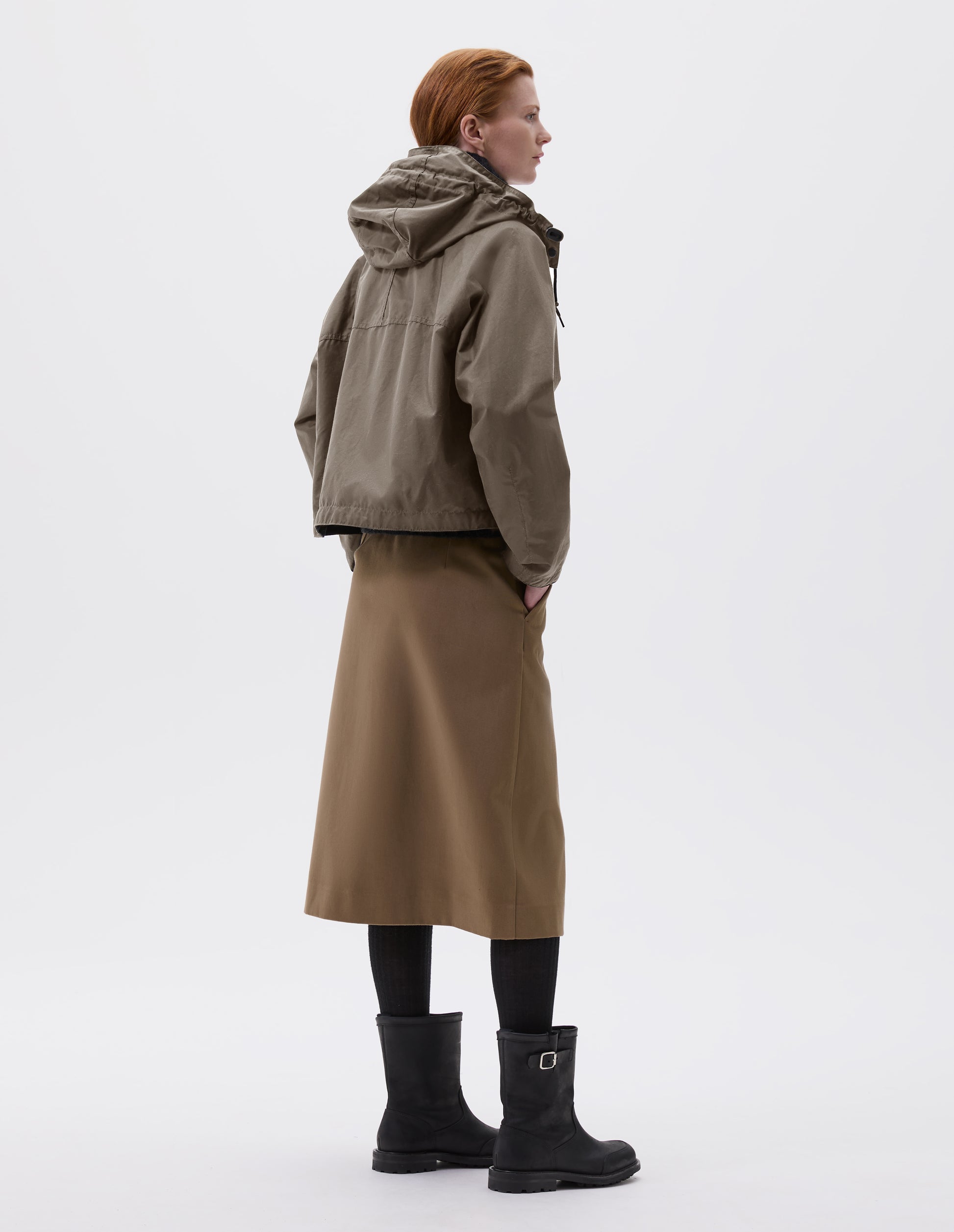 Full body image, back view of model wearing boxy dark beige jacket with raglan sleeve and hood with wire adjustable peak. Styled with beige midi skirt, black tights and black biker boots.