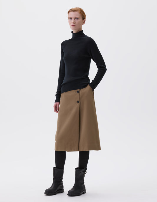 Full body image, front view of model wearing black fine knit merino wool jumper, with high neck, raglan sleeve and cast off edging on neck, cuffs and hem. Styled with brown skirt, black tights and black leather boots.