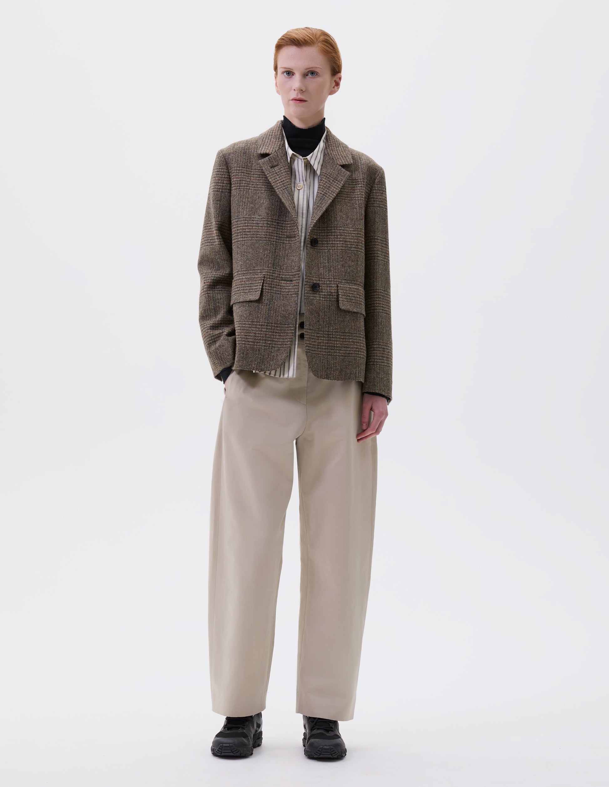 Full body image, front view of model wearing green-brown glencheck blazer with horn buttons on lapel and cuffs and flap patch pockets. Styled over black turtleneck and beige striped shirt and beige trousers with black trainers.