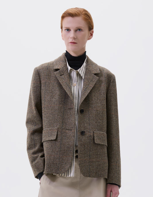 Cropped image, front view of model wearing green-brown glencheck blazer with horn buttons on lapel and cuffs and flap patch pockets. Styled over black turtleneck and beige striped shirt and beige trousers.