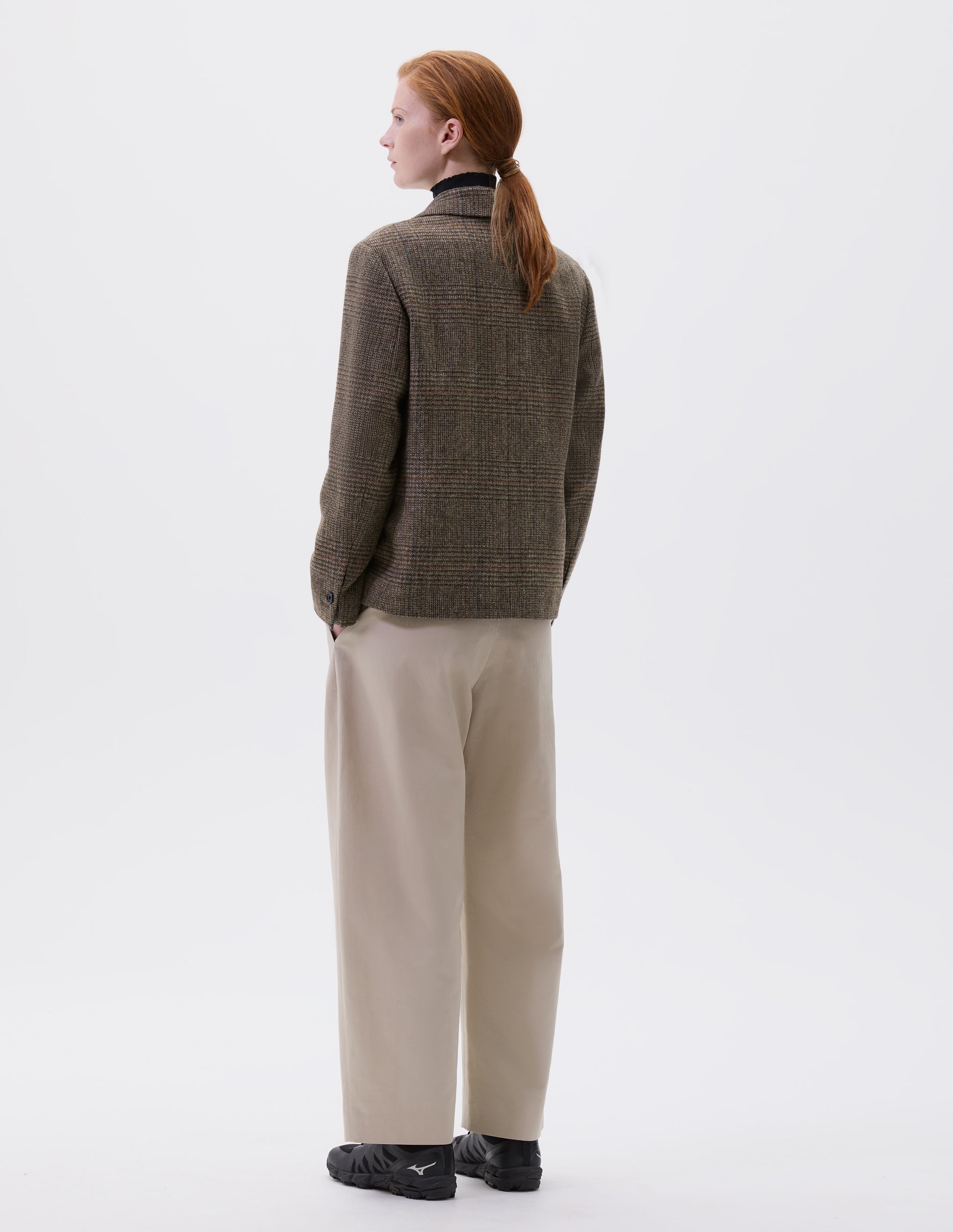 Full body image, back view of model wearing green-brown glencheck blazer with horn buttons on cuffs. Styled over black turtleneck and beige trousers with black trainers.
