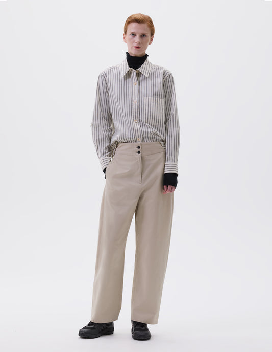 Full body image, front view of model wearing beige, grey and black striped shirt with raised placket and mother of pearl buttons, patch pocket on chest, styled over dark grey turtleneck and beige trousers with side side pockets and horn button fastening, and black trainers. 