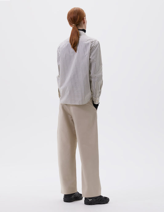 Full body image, back view of model wearing beige, grey and black shirt with mother of pearl buttons on cuffs and hanger loop detail on back yoke, styled over dark grey turtleneck and beige trousers with side pockets and black trainers. 