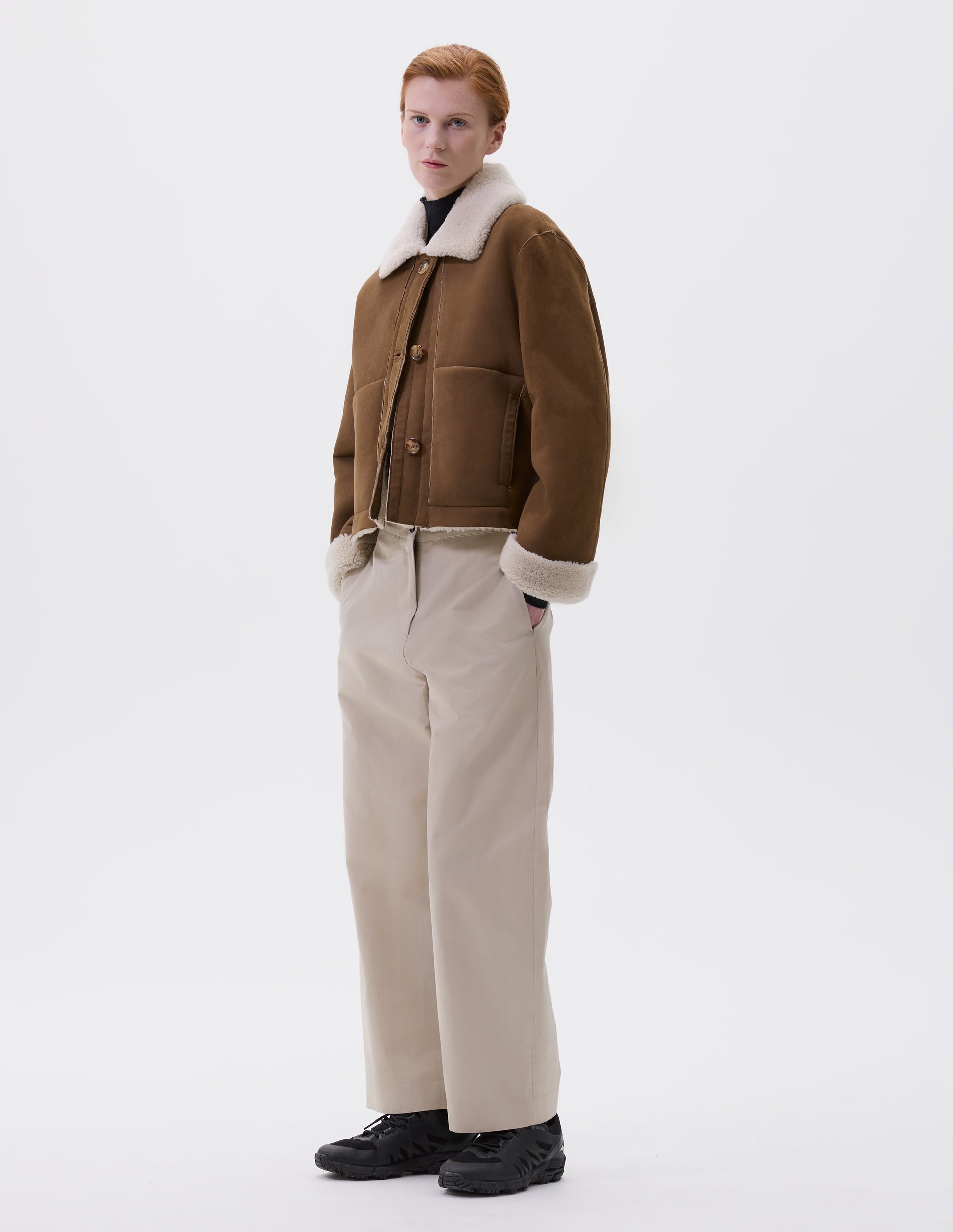 Full body image, front view of model wearing tan and ecru shearling cropped jacket, with button tab fastening collar, turn back cuffs, side welt pockets and raw edge finishing on seams. Styled over black high neck jumper, beige trousers and black trainers.