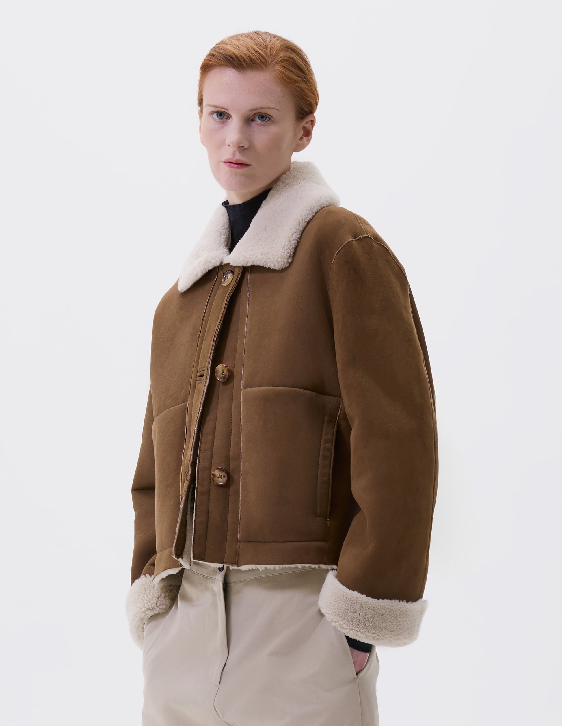 Cropped body image, front view of model wearing tan and ecru shearling cropped jacket, with button tab fastening collar, turn back cuffs, side welt pockets and raw edge finishing on seams. Styled over black high neck jumper, with beige trousers.