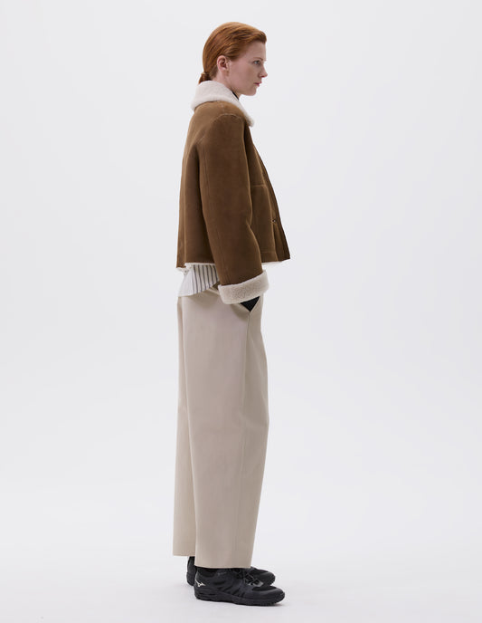 Full body image, side view of model wearing tan and ecru shearling cropped jacket, with turn back cuffs, side welt pockets and raw edge finishing on seams. Styled over striped white and black shirt, beige trousers and black trainers.