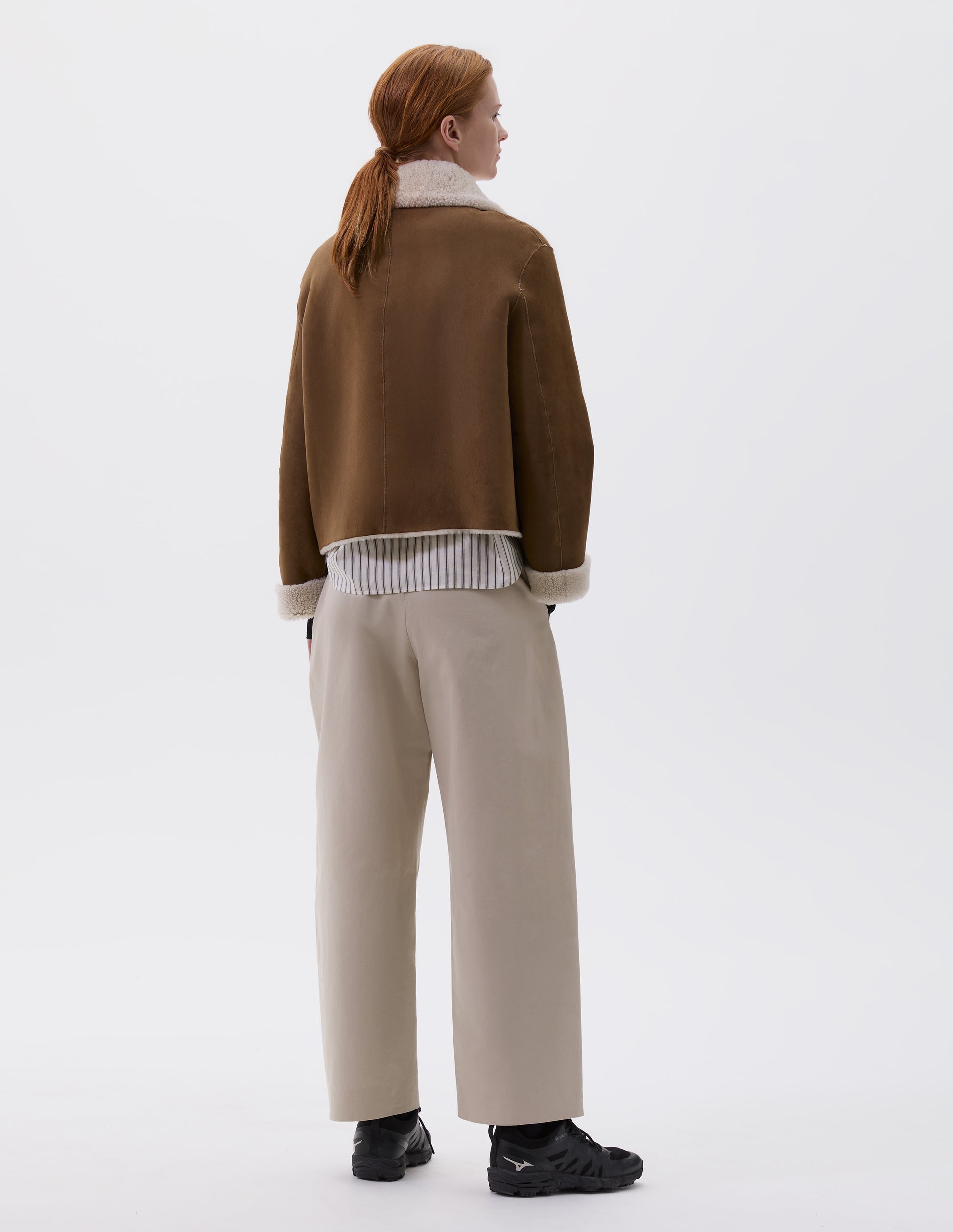 Full body image, back view of model wearing tan and ecru shearling cropped jacket, with turn back cuffs and raw edge finishing on seams. Styled over striped white and black shirt, beige trousers and black trainers.