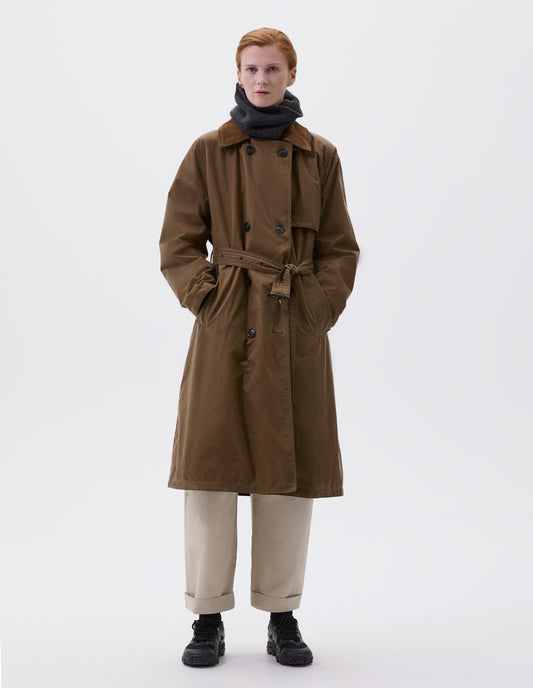 Full body image, front view of model wearing light brown trench coat with corduroy collar and horn buttons, leather buckle belt and deep welt slant pockets. Styled with dark grey neck warmer, beige trousers, black socks and black trainers.