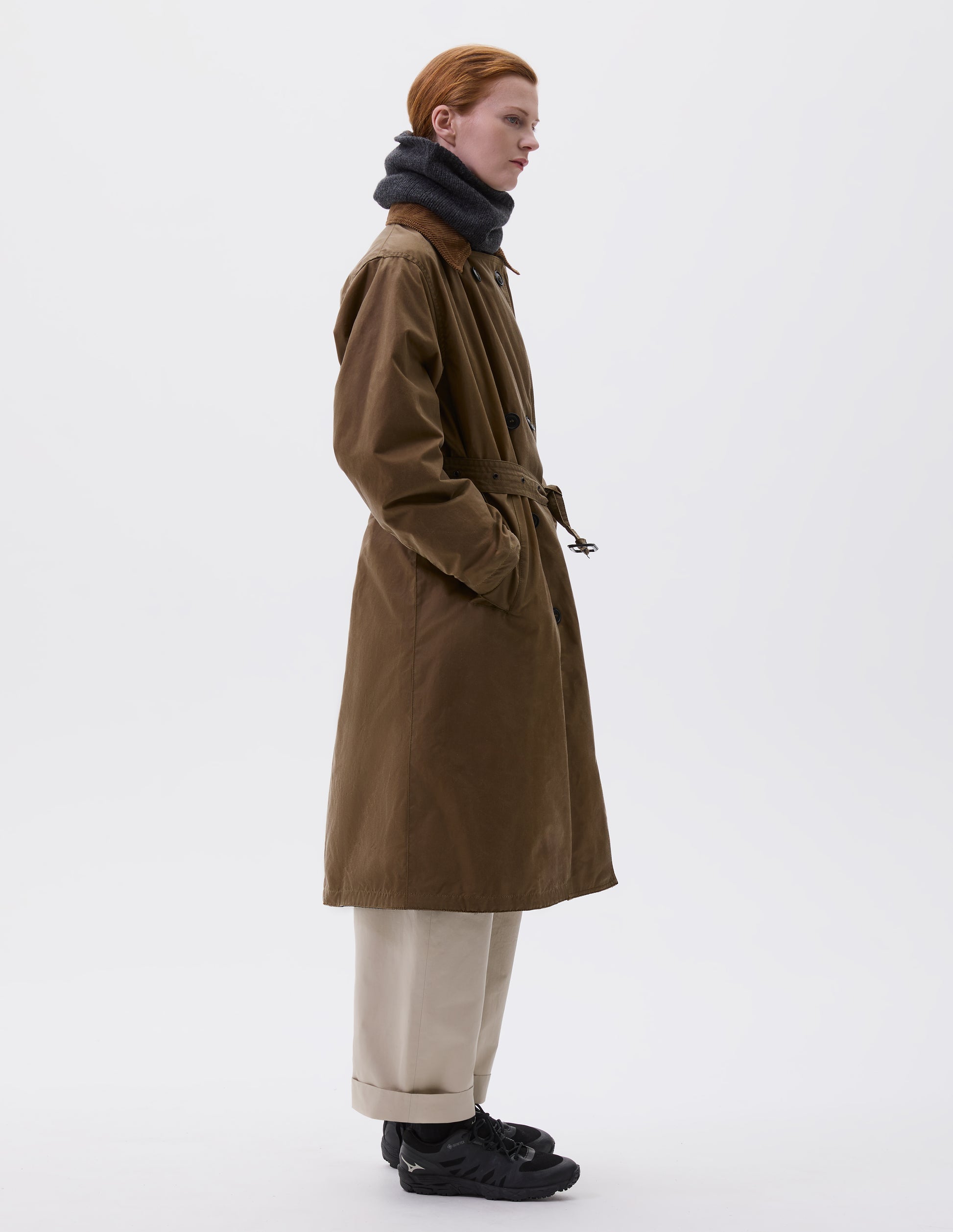 Full body image, side view of model wearing light brown trench coat with corduroy collar and horn buttons, leather buckle belt and deep welt slant pockets. Styled with dark grey neck warmer, beige trousers, black socks and black trainers.