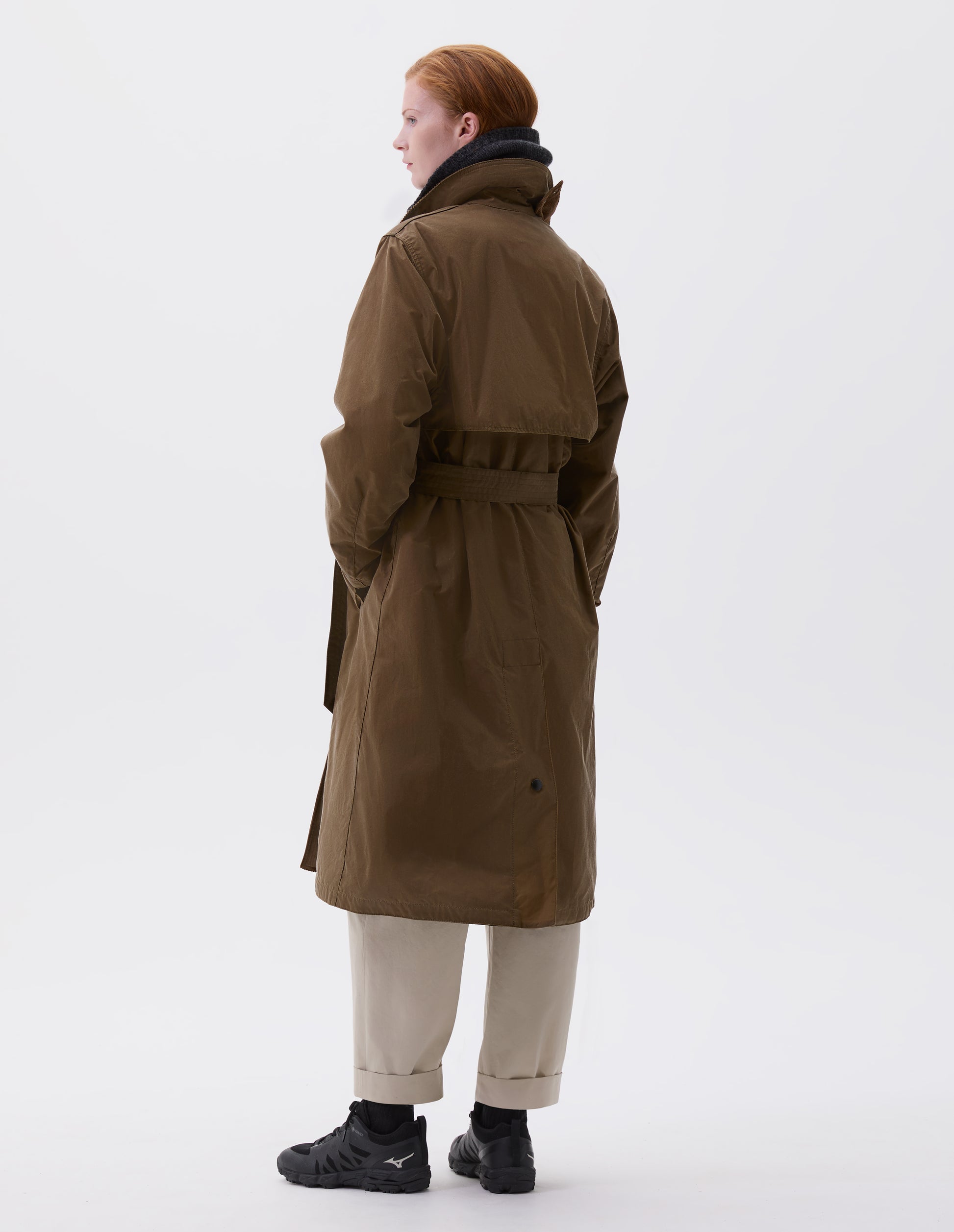 Full body image, back view of model wearin light brown trench coat with corduroy collar,  belt, storm flap and press stud fastening deep centre vent. Styled with dark grey neck warmer, beige trousers, black socks and black trainers.