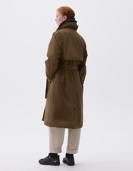 Full body image, back view of model wearin light brown trench coat with corduroy collar,  belt, storm flap and press stud fastening deep centre vent. Styled with dark grey neck warmer, beige trousers, black socks and black trainers.