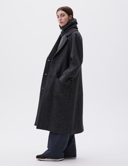 Full body image, side view of model wearing charcoal grey heavy wool longline overcoat with raglan sleeve, button fastening lapel and deep flap patch pockets. Styled with grey knitted vest, navy pinstripe trousers and black and brown trainers.