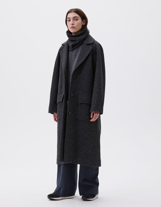 Full body image, front view of model wearing charcoal grey heavy wool longline overcoat with raglan sleeve, button fastening lapel and deep flap patch pockets. Styled with grey knitted vest, navy pinstripe trousers and black and brown trainers.