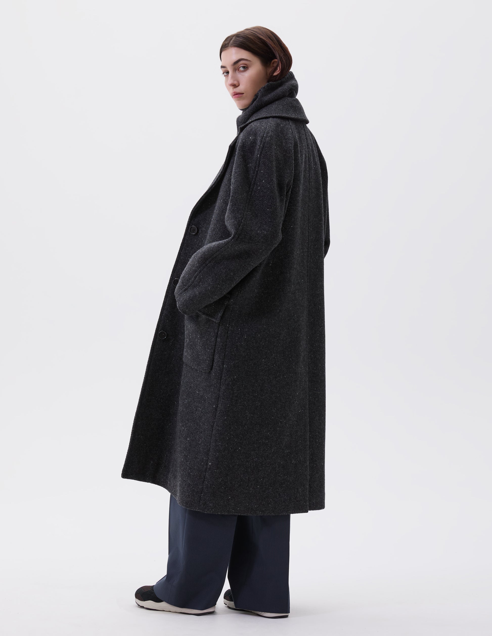 Full body image, back view of model wearing charcoal grey heavy wool longline overcoat with raglan sleeve and back centre vent. Styled with navy pinstripe trousers and black and brown trainers. 