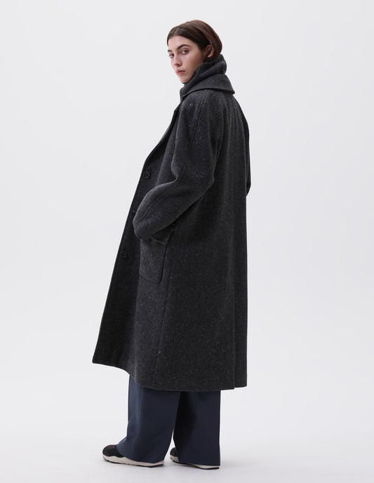 Full body image, back view of model wearing charcoal grey heavy wool longline overcoat with raglan sleeve and back centre vent. Styled with navy pinstripe trousers and black and brown trainers. 