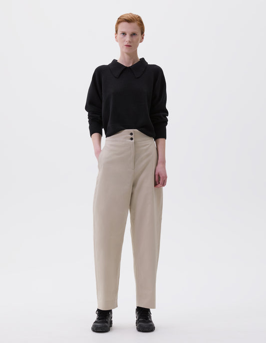 Full body image, front view of model wearing beige relaxed high waisted trousers with black horn buttons and side pockets. Styled with black collared jumper and black trainers.