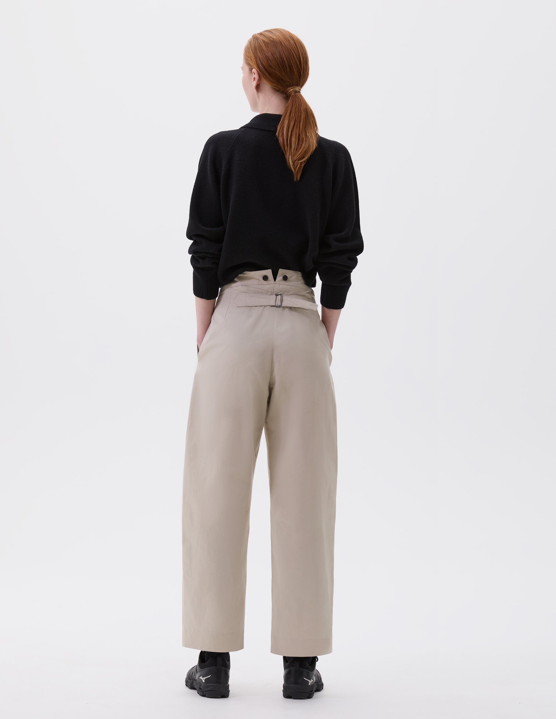 Full body image, back view of model wearing beige relaxed high waisted trousers with double black horn button details on waistband and cinch back. Styled with black collared jumper and black trainers.